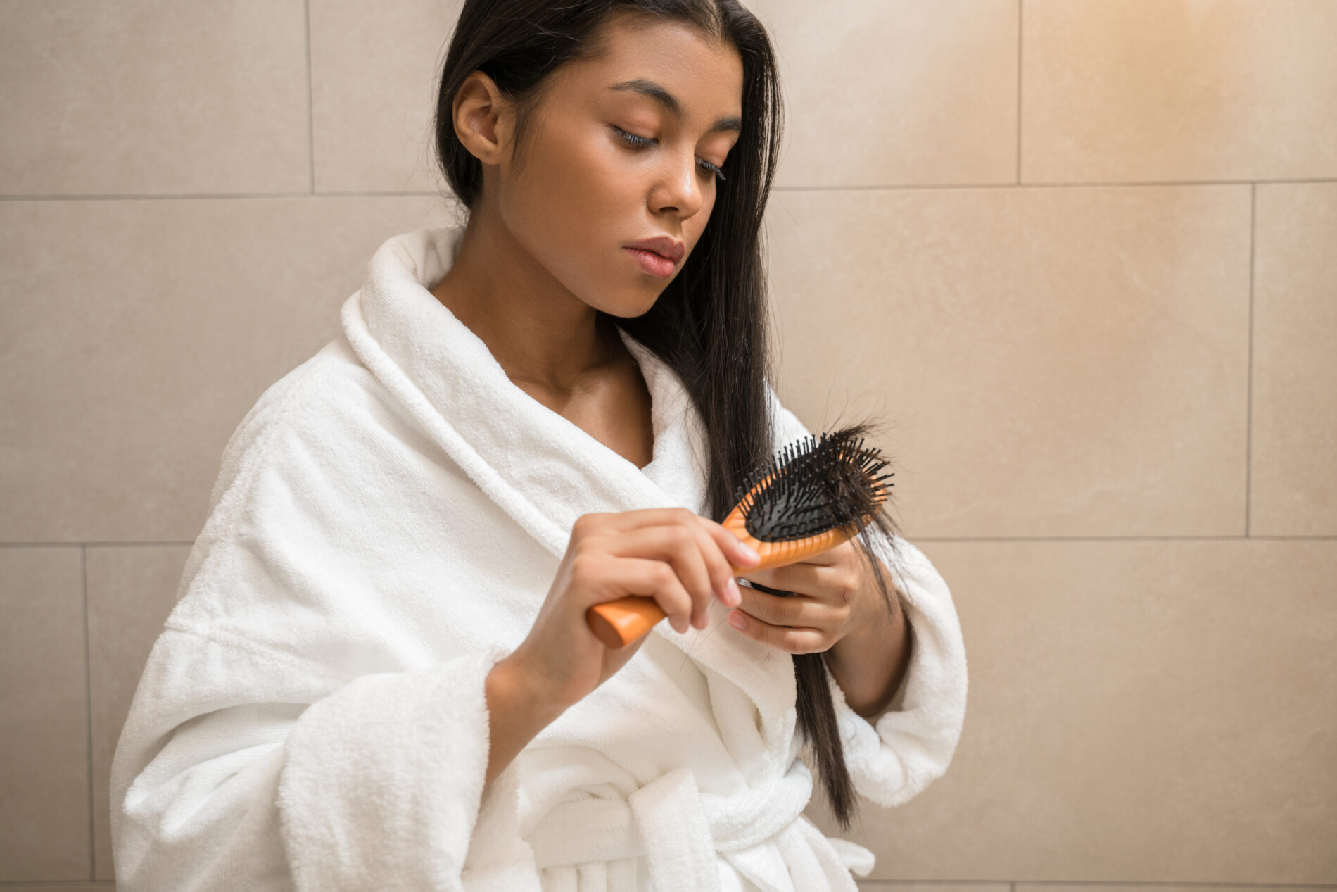 Em um banheiro está uma mulher negra, vestindo roupão branco, e penteando as pontas dos cabelos castanhos escuros e lisos, com uma escova cor de madeira clara; representando a reestruturação capilar.