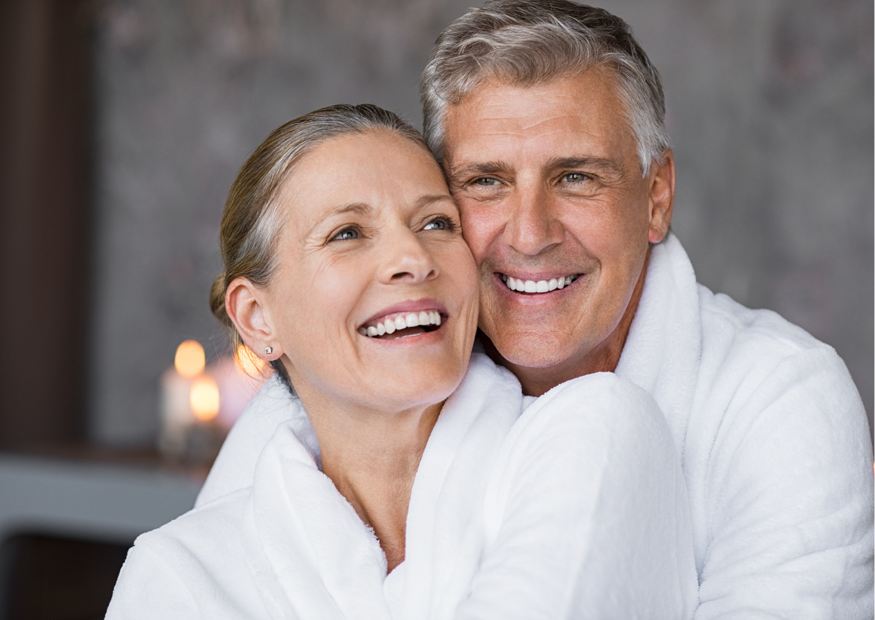 Em destaque, um casal de homem e mulher branca, ambos com cabelos claros e grisalhos, estão vestindo roupões da cor branca e abraçados e sorrindo.